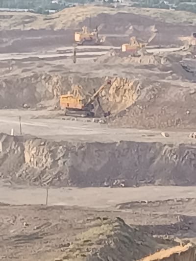 The image depicts a quarry or open-pit mine, likely located in Almalyk, Uzbekistan, given the industrial context often associated with the region. The scene is characterized by large-scale excavation and heavy machinery. In the foreground, a section of the quarry wall is visible, showing layered rock and soil. Further back, multiple excavators and possibly other mining equipment are at work. One prominent yellow excavator is positioned on a lower level of the quarry, its arm extended as if in the process of digging or moving material. Other yellow pieces of heavy machinery are scattered across various levels of the pit, suggesting ongoing mining operations. The environment is rugged and barren, with dirt, rock, and sparsely vegetated areas visible. The overall impression is one of an active industrial site dedicated to resource extraction.