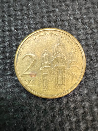 A gold-colored coin with the number "2" prominently displayed is shown on a dark, textured background. The coin has Cyrillic writing around its circumference, which translates to "2 DINARA". An image of a church with domes and a cross is visible on the coin, along with the year "2009". There is a small reddish mark on the coin near the "2". The coin is positioned slightly off-center, with the background appearing to be a woven fabric. There is no information about the city of Mejaniga, Italy in the image.