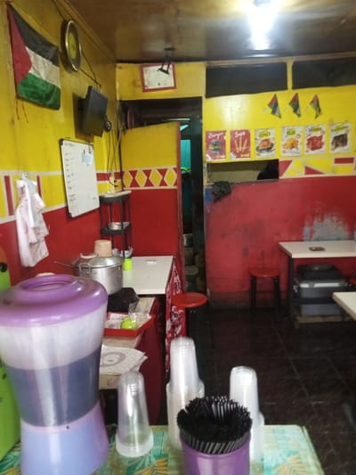 The image depicts the interior of a food establishment located in Cipendawa, Indonesia. The walls are horizontally divided, with the upper section painted bright yellow and the lower section red, separated by a red and white zigzag pattern. The ceiling is a dark, possibly wooden, surface, and the floor is covered with dark tiles.
In the foreground, a large, two-tone purple and translucent plastic beverage dispenser filled with a dark liquid rests on a table covered with a light-colored floral tablecloth. Adjacent to it are stacks of clear plastic cups and a purple container holding numerous black plastic straws.
To the left mid-ground, a white rectangular whiteboard with writing is mounted on the yellow wall. Below it hangs a white plastic bag containing wrapped items. A multi-tiered metal stand holds a small green cup on its top shelf, positioned next to a white rectangular counter. On this counter, a large lidded metal pot with a wooden handle is visible, along with another small green cup and a red plastic tray containing a green object and wrapped items. Above this, a flat-screen television is mounted on the yellow wall, and a Palestinian flag is displayed in the upper left corner. A large, circular, gold-colored object hangs nearby.
In the mid-ground right, a red stool with a black base is present. A serving counter opening is cut into the red wall. Above this opening, five rectangular posters advertise food items: "Burger (12.000)", "Sosis (20.000)", "Ayam Geprek", "Keju", "Daimono", and "Blackpepper". Three small flags resembling the Palestinian flag are mounted above these posters. A person's head and upper torso are partially visible behind the serving counter opening, facing right.
Towards the background, a doorway leads into a dimly lit area with a visible green-blue wall. A framed document is affixed to the yellow wall near this doorway. To the right of the doorway, two white rectangular tables with black bases are positioned, along with two red stools with black bases. A dark container is visible beneath the rightmost table. An overhead light fixture illuminates the scene.