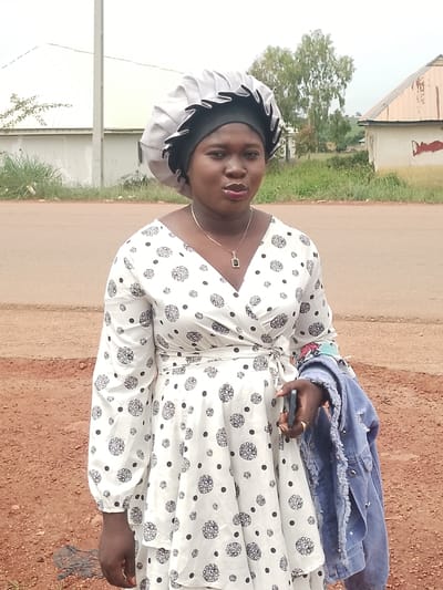 A medium shot shows a young Black woman standing outdoors in front of a road. She is wearing a black headscarf, a grey satin bonnet over it, and a white and black polka-dotted wrap dress. Around her neck, she wears a thin gold chain with a small rectangular pendant. Her left hand is holding a light blue denim jacket by its sleeve. Her right hand is bent at the elbow and rests near her hip, holding a smartphone. The woman looks towards the camera with a neutral expression and parted lips.

The setting appears to be a rural or semi-rural area in Agwala, Nigeria. Behind the woman, there is a paved road, and then a patch of reddish-brown dirt. Beyond the dirt, there is a line of sparse green grass and vegetation, followed by a road. In the background, there are a few buildings, one with a white facade and a light brown, sloped roof, and another larger white structure. A few trees are visible in the distance. The sky is overcast, suggesting a cloudy or possibly rainy day. The lighting is diffused, indicating it is likely daytime. There is no visible text in the image. The overall mood is casual and candid.