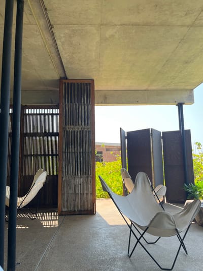 The image shows a covered patio with concrete floors and a concrete ceiling. The patio is located in a building with a wooden slatted wall on the left side of the frame. There are two butterfly chairs, with white canvas seats and black frames. The chairs are facing outwards from the patio. The patio is enclosed by a dark-colored metal slatted wall on the right side of the frame. There is a partially visible brick building in the background. There is greenery and foliage visible outside the walls and on the ground. The coordinates are 15.939° N, 97.279° W, which correspond to San Isidro Llano Grande, Mexico.