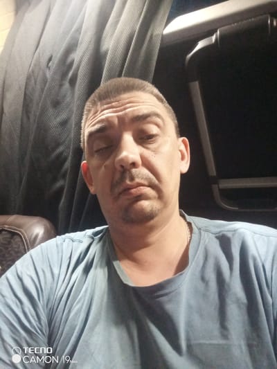 The image displays a close-up, head-and-shoulders portrait of an adult male, estimated to be 40-50 years old, located in the city of Ильинское-Усово, Russia. The subject has short-cropped light brown hair with a receding hairline and short facial hair consisting of a goatee and stubble. His forehead exhibits several horizontal wrinkles, and his expression is neutral. He is wearing a light blue, short-sleeved crew-neck t-shirt, and a thin, light-colored metal chain is visible around his neck.
The background elements suggest an interior environment, consistent with a vehicle's cabin. To the lower left, a dark brown, upholstered surface with a stitched pattern is visible, likely a seat or headrest. Behind the subject, towards the upper left, there is a dark grey, finely textured fabric with distinct vertical pleats, indicative of a curtain or interior panel. To the upper right, a segment of a light-colored horizontal trim piece is present, below which a dark, curved handle or latch mechanism is visible. The area immediately to the right of the handle is dark, suggesting an unlit compartment or window at night. The lighting in the scene is subdued. A 'TECNO CAMON 19 Neo' watermark is embedded in the bottom left corner of the photograph.
