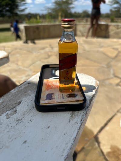 An outdoor scene in Olkaria ward, Kenya, under bright daylight. In the foreground, a miniature glass bottle of Johnnie Walker Red Label Scotch Whisky, partially filled with amber liquid and secured with a red and gold screw cap, stands upright. It is positioned inside a black smartphone case, which contains a single Argentinian 1000 Pesos banknote featuring a Hornero bird, "MIL PESOS," and "José de San Martín" text. These objects rest on a weathered, off-white surface, possibly a painted wooden railing or concrete edge. The ground in the midground is paved with light-colored, irregularly shaped stones. A low, light-colored concrete or stone barrier or bench is visible further back. The background shows two out-of-focus individuals: one in a blue garment to the left and another with dark skin wearing a dark top and shorts to the right. Sparse trees and shrubs are present in the distant background under a clear sky. Distinct shadows indicate direct sunlight.