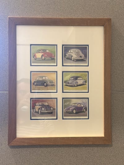 A framed collection of six vintage Volkswagen Beetle illustrations is displayed on a textured grey wall. The frame is made of wood and has a light brown, matte finish. The illustrations are arranged in two columns and three rows, with each illustration featuring a different model and year of the Volkswagen Beetle, along with its designation printed below.
From top left to bottom right, the illustrations depict:
- A red and yellow 1949 Hebmüller Cabriolet.
- A light grey 1951 Export Beetle.
- A dark green 1952 Beetle.
- A light grey 1954 Karmann Cabriolet.
- A black and cream 1959 Karmann Cabriolet.
- A purple 1966 Beetle 1300.
The wall behind the frame is a speckled grey, possibly concrete or stone. The lighting appears to be from above and slightly to the left, casting subtle shadows. There are no people or animals present in the image. The scene appears to be an indoor setting, likely a home or a commercial space in Ouahat Sidi Brahim, Morocco.