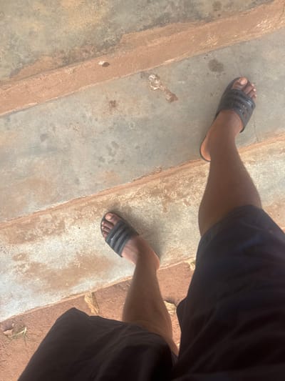 A view from above shows legs and feet on a stone-like surface with a textured or uneven border. The person wears black Adidas-style slides. The legs are angled, with one foot positioned higher in the frame than the other. The person is wearing dark shorts. The overall perspective suggests a casual or candid snapshot of someone standing on a rough or outdoor surface. The lighting appears to be natural.