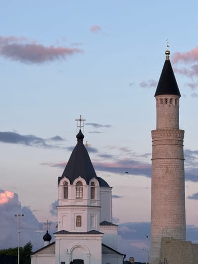 This is a vertical, low-angle, eye-level shot of a minaret and part of a mosque in Bolgar, Russia. The sky is a pale blue with wispy clouds tinged with pink and purple, suggesting either dawn or dusk. The minaret is tall and cylindrical, constructed from light-colored brick with decorative bands and an arched balcony near its pointed, dark roof topped by a crescent moon. To the right and lower in the frame, a white mosque building is visible, featuring a dark, conical roof with arched windows and a cross at its peak. The architectural style suggests a blend of Islamic and Christian influences, possibly indicating historical context or religious coexistence in the area. A small dark bird is in flight in the sky between the two structures. The overall mood is serene and atmospheric, enhanced by the soft lighting of the twilight sky. There is no visible text.