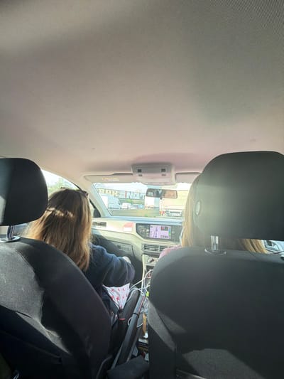 The image captures the interior of a vehicle, viewed from a rear passenger seat, looking forward onto a multi-lane highway near Aulnay-sous-Bois, France. The vehicle's interior features black fabric front seat headrests in the foreground and a light grey fabric headliner.

Two individuals occupy the front seats. The driver, seated on the left, has long, light brown hair, with sunglasses positioned on her head, and is wearing a dark blue long-sleeved garment. Her hands are visible near the steering wheel. The front passenger's head, with light brown hair, is visible on the right. The driver's eyes and forehead are discernible in the reflection of the rear-view mirror.

The dashboard, dark in color, features a central infotainment screen displaying a navigation map. The center console area between the front seats contains multiple white cables and a white fabric item patterned with red stars.

Through the windshield, a multi-lane highway is visible, populated by moderate to heavy traffic including several white semi-trailer trucks and passenger vehicles, suggesting either congestion or active travel. Spanning the highway above the traffic is a large grey gantry sign with partially visible white text, notably the phrase "BAR NOT," confirming the location as the A3 highway in the vicinity of Aulnay-sous-Bois, France. Structures and vegetation are visible in the background beyond the highway.