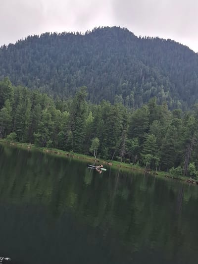 The image depicts a tranquil natural scene at Lake Vodopadnoe, located near the village of Vydrino, Russia. A dense forest of coniferous trees covers a steep mountain slope that forms the backdrop. The trees are a deep green, creating a rich texture against the overcast sky. In the foreground, the calm surface of the lake reflects the dark green of the forest and the muted colors of the sky. A small raft or paddleboard with two individuals is visible on the water, positioned closer to the treeline. The overall impression is one of serenity and natural beauty.