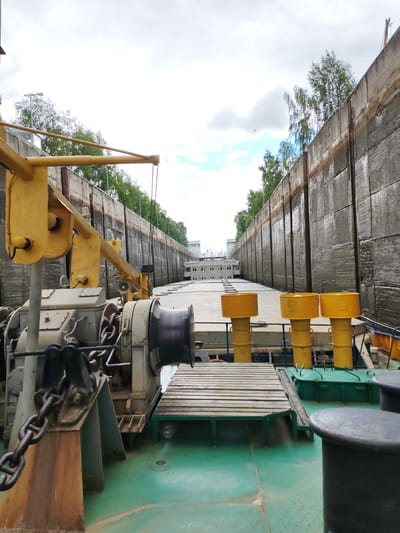 The image shows a view from the deck of a barge or ship, looking forward into a concrete lock chamber. On the left side of the foreground, there is a large yellow crane structure with chains and an anchor winch. The barge is positioned in the lock, with tall, textured concrete walls rising on either side. Along the right side of the lock, several yellow, cylindrical objects with funnel-shaped tops are visible, mounted on poles. Further ahead, the bow of another vessel is visible, and beyond that, buildings can be seen in the distance. The sky is overcast with some patches of blue. The scene depicts a segment of the White Sea-Baltic Canal, likely within the city of Vytegra, Russia, a key location on the waterway.