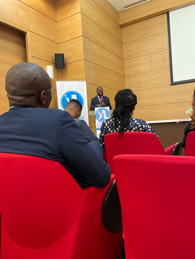 The image depicts an indoor conference or presentation taking place in what appears to be an auditorium or meeting room. A man, dressed in a dark suit, light shirt, and tie, stands at a podium in the mid-ground, speaking into a microphone. He is positioned in front of a blue and white banner that features a logo and partially visible text, including "AL", "CENTER", and "NATIONAL LEADE".
In the foreground, two individuals are seated in red, upholstered chairs, with their backs to the viewer. The closest person is a bald man wearing a dark suit jacket, with glasses resting on the back of his head. Behind him, another person with long dreadlocks is visible, wearing a dark patterned top. A third person with lighter hair is partially visible on the far right. The attendees are facing the speaker, indicating they are part of the audience.
The room's walls are covered with light wooden paneling. A large white projection screen is visible on the left wall, along with a black wall-mounted speaker or device below it. The rows of red chairs suggest a formal seating arrangement for an audience. The lighting is consistent with an indoor daytime event, likely a seminar, speech, or workshop. The atmosphere appears attentive and engaged.
