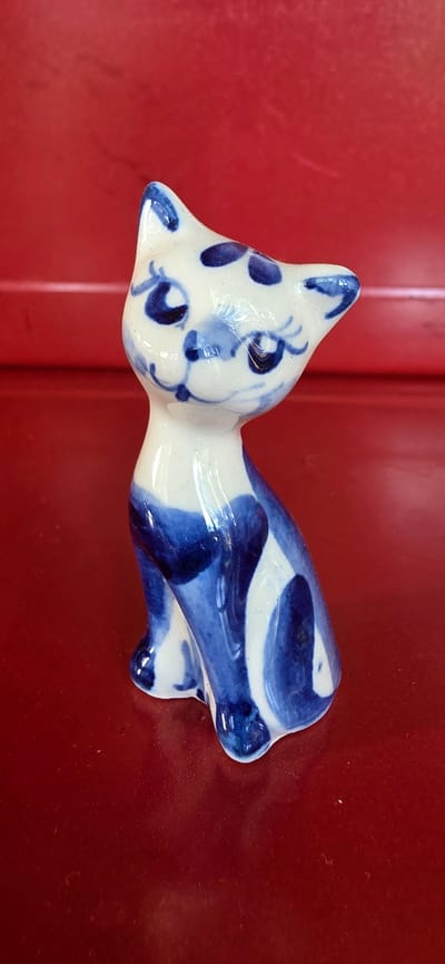 A white ceramic cat figurine with blue floral patterns is placed on a shiny red surface. The figurine is sitting and facing to the left, with its head slightly tilted upwards. The background is a blurred red surface with a slight texture. The image does not contain any information that would allow for localization in the city of Электротехник Снт, Russia.