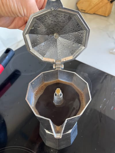 This image depicts a silver-colored, octagonal Moka pot with its lid open, revealing freshly brewed dark coffee inside the upper chamber. The coffee displays a light brown foam ring along its perimeter and appears to be actively bubbling from the central conduit. The interior of the hinged lid is covered in numerous condensation droplets, indicative of recent heat and steam. A human hand, positioned in the upper left, holds the lid in an open position. The Moka pot rests on a black induction or ceramic stovetop, with a red and black utensil handle partially visible on the left side of the stovetop. A light-colored countertop or wall is present in the blurred background, along with a indistinct wooden object. The scene is located in a kitchen in La Barra, Uruguay.