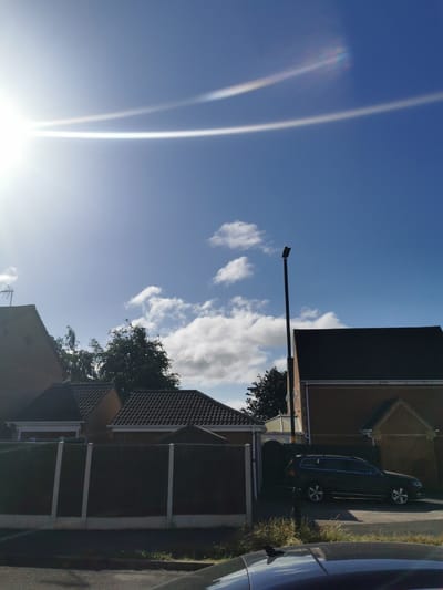 In Ripon, United Kingdom, an outdoor scene unfolds under a bright blue sky with scattered white clouds. The sun is high and bright, creating lens flares that streak across the upper portion of the image. In the foreground, a dark-colored vehicle is partially visible, its roof and a shark fin antenna suggesting it is parked or in motion on a road. Behind the vehicle, a dark fence with white concrete posts delineates a property boundary.
Beyond the fence, several residential buildings are visible. To the left, the upper portion of a brick house with a red-tiled roof and a television antenna is seen. Adjacent to it is another building with a similar red-tiled roof, possibly a garage or extension. Further to the right, a larger brick house with a dark-tiled roof dominates the view. A tall, black lamppost stands in front of this house. Trees with dark green foliage are interspersed between the buildings and behind the fence. The overall impression is a suburban residential area on a sunny day.
