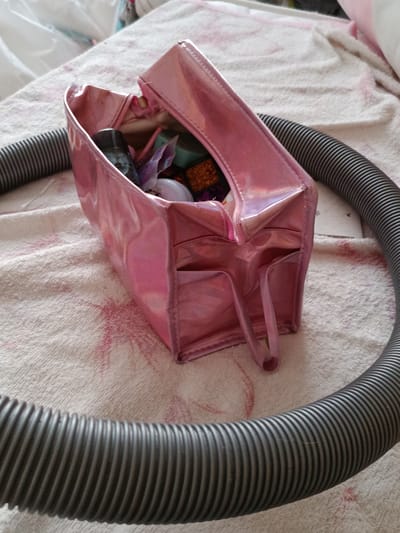 A pink, iridescent makeup bag sits open on a textured, light-colored fabric, possibly a towel or blanket. The bag is filled with various small items, including what appears to be makeup products and colorful trinkets. A ribbed, dark gray flexible hose is looped around the makeup bag, forming a large circle. The fabric has a subtle pink pattern. The scene is indoors, with a blurred background suggesting other soft furnishings. There are no people visible in the image. The location in Novotitarovskaya, Russia, is not discernible from the visual information provided.
