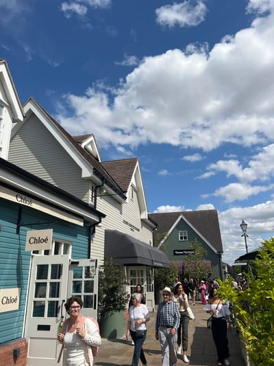 The image depicts a sunlit outdoor retail environment identified as Bicester Village in Bicester, Cherwell District, United Kingdom. The scene presents a paved pedestrian walkway flanked by boutique buildings designed in a village style with gabled roofs and varying color schemes.
On the left, a light blue-green building with white trim houses a "Chloé" storefront, marked by two white signs bearing the brand name and a white multi-paned front door. Adjacent to it stands a light grey building with a brown roof and a dark retractable awning, identifiable as "Golden Goose Deluxe Brand" by its partial signage. Further down the pathway on the right, a dark green building with a gabled roof and a single white window hosts a "Vivienne Westwood" outlet, with its name visible below the window.
Approximately ten to fifteen individuals are present on the walkway, moving primarily from left to right or towards the camera. In the foreground, a woman with dark curly hair and glasses, wearing a white tank top and a light-colored cardigan, is seen smiling and holding a light-colored bottle in her left hand. Other visible pedestrians include an older woman in a white t-shirt, an older man in a plaid shirt and a straw hat, and a woman in a white off-the-shoulder top. The environment features large potted plants near the storefronts, green shrubbery along the right side of the path, and a black ornamental street lamp. The sky is clear blue with scattered white cumulus clouds.