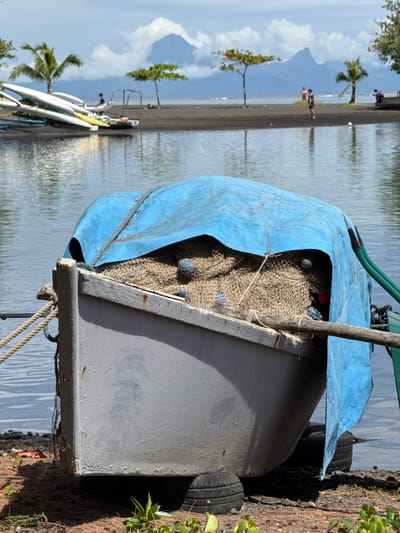 A light gray boat, partially covered by a weathered blue tarp, is beached on dark ground in the foreground. Beneath the tarp, tan fishing nets with visible blue and red floats are stored inside the boat. A thick rope is tied to the boat's bow. Green foliage grows in the immediate foreground. Beyond the boat, a calm body of dark water reflects the sky. Across the water, a dark, possibly volcanic, sand beach is visible. On this beach, located in Māhina, France, a stack of white and yellow kayaks or canoes is present on the left. Three distinct small trees and two palm trees are scattered along the beach. Several individuals are present: one stands centrally, one is partially visible to the far right, and another appears to be reclining on the far left. In the distant background, towering, jagged mountains are partially obscured by white clouds against a blue sky.