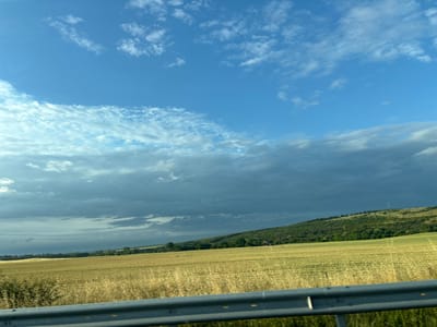The image depicts a rural landscape with a field of dry grass or crops in the foreground, extending towards a tree-covered hill in the background. A grey metal guardrail runs along the bottom of the frame, suggesting the photo was taken from a moving vehicle. The sky is partly cloudy with patches of blue sky and darker grey clouds. The location is identified as Rupkite, Bulgaria. There are no people or animals visible in the image.