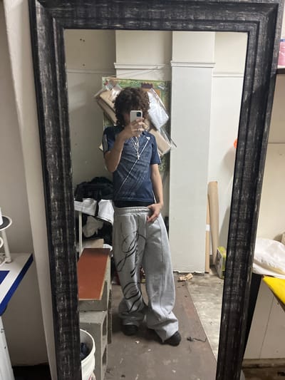 A person with curly brown hair is taking a photo of themselves in a large, distressed black-framed mirror. The person is wearing a blue plaid shirt and gray sweatpants with black markings. The environment reflected in the mirror includes a white cabinet, a brown baseball bat leaning against the wall, and a concrete floor. In the lower right of the reflection, there are cinder blocks and a white bucket. It is difficult to provide the precise location in Bahía Blanca, Argentina, without more context or landmarks visible in the image.