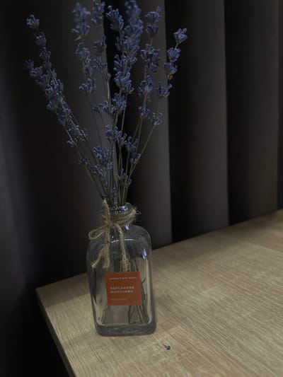 A still life image shows a glass bottle containing dried lavender sprigs, standing on a light brown wooden surface. The bottle is tied with jute twine. An orange label on the bottle reads "АРОМАТ ДЛЯ ДОМУ" (Home Aroma) and below it, in larger Cyrillic text, "ПЕРСИКОВЕ МОРОЗИВО" (Peach Ice Cream). Underneath this, it states "100 мл (ml)". The background consists of dark grey vertical curtains. A single dried lavender flower lies on the wooden surface in the lower right portion of the image. The scene is indoors. The location is Peremyshliany, Ukraine.