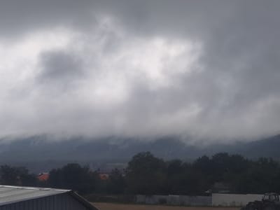 The image depicts a cloudy, overcast day in Markhua, Russia. In the foreground, the metal roof of a structure is visible. Beyond this, a low fence runs horizontally across the scene. In the middle ground, a dense line of dark green trees obscures most of the lower portion of the distant hills. Several rooftops, some with orange tiles, peek through the trees. A tractor is partially visible on the right side of the frame. The background is dominated by a mountain range shrouded in thick, low-lying clouds, with the sky above filled with further layers of gray clouds.