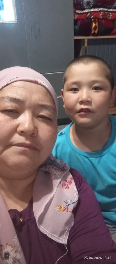 In a close-up, indoor selfie taken in Chon-Kara, Kyrgyzstan, a woman wearing a pink headscarf and a purple shirt is partially visible on the left. Her left eye is closed, and she is looking towards the camera. To her right, a young boy with short hair and a blue t-shirt is facing the camera. The background is indistinct, appearing to be a room with a gray wall on the left and stacked colorful textiles on the right. A timestamp in the bottom right corner indicates "23.06.2026 18:15".