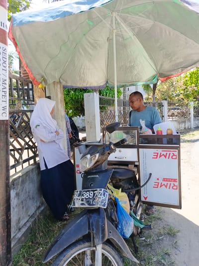 A man and a young woman are at a roadside stall under a large umbrella in Meuliek, Indonesia. The man, wearing a blue t-shirt, is tending to the stall, which is equipped with a motorcycle. The stall displays food items and condiments. The young woman, wearing a white hijab and school uniform, stands with her arms crossed, facing away from the stall. The surrounding environment includes a concrete wall, a metal fence, and lush green trees. The ground is a mix of dirt and grass. The scene is captured on a bright, sunny day.