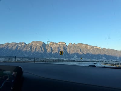 A landscape viewed from a car shows a highway with yellow guardrails and signage leading to mountains. The mountains are illuminated by the sun, casting shadows. The sky is clear and blue. The interior of the car is visible at the top of the frame, including a screen. The image appears to be taken through the windshield.
