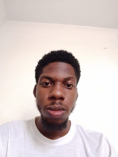 This is a close-up, head-and-shoulders selfie of a young Black man in Abeere, Nigeria. He is looking directly at the camera with a neutral expression. He has dark, short, curly hair and a short beard. He is wearing a plain white t-shirt. The background is a plain, light-colored wall. The lighting appears to be natural, suggesting it is daytime. The overall impression is casual and informal. There is no visible text in the image.