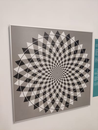 The image shows a framed optical illusion artwork on a white wall. The artwork itself is a circular pattern of black and white triangles that create a sense of spiraling motion. The background of the artwork is a textured gray. To the right of the artwork, a portion of a teal informational panel is visible, containing text in both Greek and English. The Greek text at the top discusses a spiral and "real circles," while the English text below, under the heading "When the chessboard illusion appears," explains that the image is about a "false impression" and "smoothness" created by the "light and dark ground." The setting appears to be a museum or exhibition space in Thessaloniki, Greece, given the bilingual text and the display of an optical illusion. There are no people, specific time of day, or weather conditions evident.
