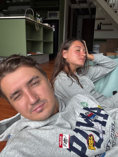 A couple, possibly on a couch, is captured in a rotated selfie. The woman, with closed eyes, rests her head. The man winks at the camera, revealing light stubble and neatly styled hair. Both are dressed in matching grey sweatshirts. The man's sweatshirt has a prominent "Puma" logo adorned with various patches and phrases such as "Don't Flinch" and "Hello My Name Is." Behind them, part of an organized home is visible with shelves, bottles, and a TV. The overall image is relaxed and intimate.