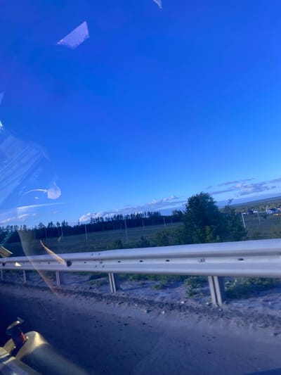 The media file depicts a scene from a moving vehicle, likely a car, looking out a side window. The perspective is tilted, with the horizon appearing to be at a 90-degree angle. The primary subjects are the sky, a guardrail, and a landscape.

The sky is a vibrant, clear blue, suggesting a bright day. A few wispy clouds are visible in the upper portion of the sky, adding some texture. In the middle ground, a line of trees runs parallel to the road, with some larger trees interspersed. Beyond the trees, there is a body of water, possibly a lake or river, and on the far bank, some buildings with dark roofs are visible, hinting at a rural or suburban settlement. A white guardrail runs along the right side of the frame, marking the edge of the road. The road surface itself appears to be gravel or dirt.

The scene suggests travel, with the blurred reflections in the window indicating motion. The lighting and clear sky point towards daytime, possibly late afternoon or early evening given the angle of the sun might be lower, though this is not definitively clear from the image. No specific activities or events are taking place, it is a snapshot of a journey.

Notable details include the reflection of the interior of the vehicle in the window, showing parts of the car's interior and a person's arm. The overall mood is calm and observational, typical of a scenic drive. No visible text is present in the image. The location context provided is Заглядовка, Russia.