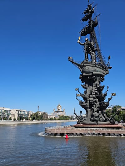 The image shows the Monument to Peter the Great in Moscow, Russia. The monument is a large bronze statue of Peter the Great standing on a ship, with several smaller ships and masts surrounding him. The statue is located on an island in the Moskva River, with the Christ the Saviour Cathedral visible in the background. The sky is clear blue, and the water is calm. There are some buildings on the riverbank to the left of the statue. The monument is intricately detailed, with many small figures and objects depicted on the ships and masts. The overall impression is one of grandeur and historical significance.