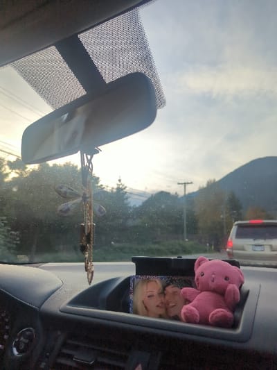 The image, captured from the interior of a vehicle, shows the dashboard and the view through the front windshield. In the immediate foreground, a rearview mirror is suspended from the windshield's top edge, with a decorative dragonfly-shaped charm featuring purple translucent wings and dangling elements hanging below it. The dashboard, dark gray in color, features air vents and a central recessed compartment. Within this compartment, a small framed photograph depicts two individuals: a blonde female and a male with dark hair and a head covering, both smiling or laughing, with the female appearing to playfully lick the male's cheek. Adjacent to the photo, on its right, a small, pink plush teddy bear sits upright.

Through the windshield, a road environment is visible. Green trees and foliage line the left side of the road and extend into the mid-distance. Utility poles and power lines are also discernible. In the right lane or shoulder, a light-colored (possibly beige or silver) SUV with illuminated taillights is present. In the background, distinct mountain ranges, consistent with the geography of Area G (Deroche/Dewdney/Sumas Mountain), Canada, rise against a partly cloudy sky. The lighting suggests either early morning or late afternoon conditions.