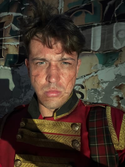 A close-up, medium shot captures a person's face and upper torso against a textured, torn background. The individual has light skin with several visible scars on their face, particularly on the left cheek and forehead. Their eyes are blue and appear to be looking directly at the camera. The person has brown hair that is slightly messy. They are wearing a red military-style jacket with gold braiding and buttons, and a green collar. A plaid fabric, predominantly red and green, is visible on the right shoulder. The background is a wall covered in torn posters or paper, with muted colors and abstract shapes, suggesting an urban or derelict environment. There is a shadow cast on the left side of the background, obscuring part of the wall. The image appears to be taken in Gollagutta, India.
