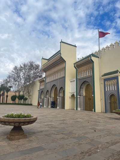 A wide-angle exterior view depicts the ornate gates of the Royal Palace of Fes in Mechouar Fes Jdid, Morocco, under a partially cloudy sky. The structure features light yellow walls accented with intricate Moroccan zellige tiling in shades of green, blue, and gold, arranged in geometric patterns around multiple arched doorways. Several large, golden-colored doors are visible, some within the arches. The building facades incorporate decorative cornices and columns, topped with green-tiled rooflines and a crenellated wall section on the far right. A reddish-maroon flag is flown from a flagpole positioned above the rightmost section of the palace wall.
In the foreground, a large, tan-colored paved plaza extends across the scene. A multi-tiered stone planter containing green foliage and white flowers is situated in the lower-left foreground. To the left of the palace gates, a tall, light-colored wall with palm trees and leafless deciduous trees extends into the background.
Several individuals are present on the plaza. Two figures, an adult in a dark coat and a child in lighter clothing, walk away from the viewer towards the left side of the scene. A small group of three to four individuals, dressed in dark clothing, stands near one of the central arched entrances, appearing to be in the process of entering or exiting. The overall lighting suggests an overcast or late afternoon setting.