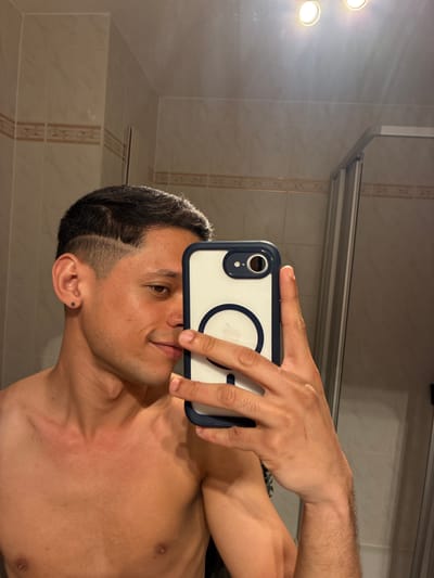 A shirtless individual is taking a selfie in a bathroom. The individual is holding a smartphone with a clear case with a MagSafe ring on the back. The bathroom has tiled walls and is illuminated by overhead lighting. The individual has short, dark hair and appears to have a piercing in their ear. There is a shower door visible in the background. There are no indications of the location being Cottbus - Chóśebuz, Germany.