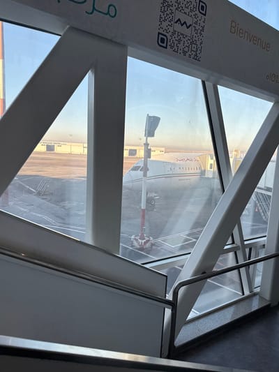 The image captures an airport tarmac scene viewed from inside a modern terminal or jet bridge in Nouaceur, Morocco. Structural elements of the building, comprising white metal beams and large glass panels, dominate the foreground and mid-left. A white staircase with silver handrails is partially visible in the lower foreground, descending to the right. Above, a white sign panel displays a black QR code with an "MW" logo, partial Arabic script, and the French word "Bienvenue."

Through the window, a white passenger aircraft, identified by text on its fuselage as a "BOEING 737-8", is parked on the tarmac. The aircraft features a distinct red stripe running along its fuselage and Arabic script above it. A mobile jet bridge is directly connected to the aircraft's forward passenger door. To the left of the aircraft, a tall pole with red and white horizontal stripes, topped by a dark, rectangular device, stands on the tarmac. White lines mark the tarmac surface.

In the background, a low, elongated white building is visible, partially labeled "LAST BAGGAGE CENTER" and "CMN." The sky is clear and blue, with a faint atmospheric haze near the horizon. The scene is illuminated by natural light, suggesting either morning or late afternoon. No individuals are discernible in the frame.
