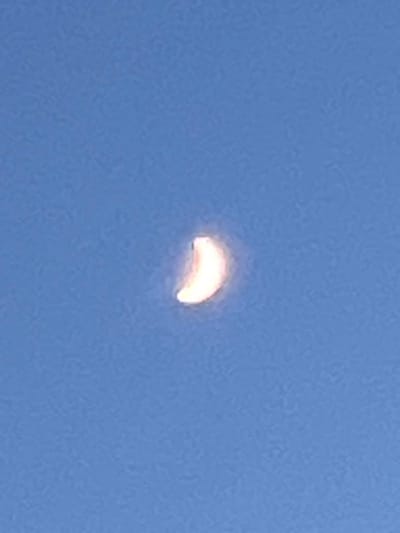 The image shows a crescent moon against a clear blue sky. The moon is illuminated and appears to have a slight reddish-orange hue. There is no indication of the city of Ilavche, Ukraine, or any other specific location within the image. The scene is a simple celestial view with no people, objects, actions, or interactions present.