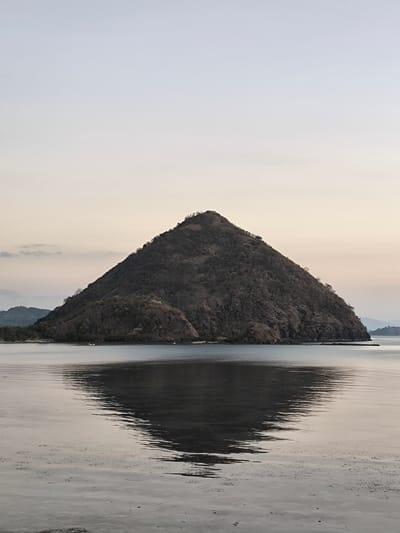 An almost perfectly conical island rises from a calm body of water, its slopes covered in dry, sparse vegetation and rocky outcrops. The island is dark brown in color, with a slightly darker summit dotted with small trees. The sky above is a pale, hazy gradient, transitioning from a light blue at the top to a soft, peachy hue near the horizon. The water in the foreground is still, reflecting the island and the sky in a distorted, rippling mirror image. In the distance, to the left, a low, hazy mountain range is visible, and a small white boat can be seen on the water near the shore of the conical island. The overall scene evokes a sense of tranquility and natural beauty, likely captured at dawn or dusk. This conical island is Kila Island, also known as Kelor Island, located in Labuan Bajo, Indonesia.