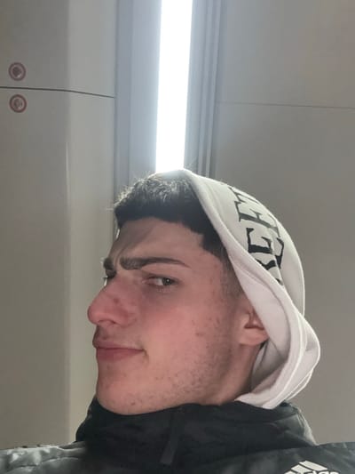 A young man with short dark hair and fair skin looks down at the camera. He has a slight smile. He is wearing a white hoodie with black lettering. Part of a dark jacket with white detailing is visible on the right. The background is a light grey surface and a light fixture. Two red circles with white smiley faces appear in the lower left corner.