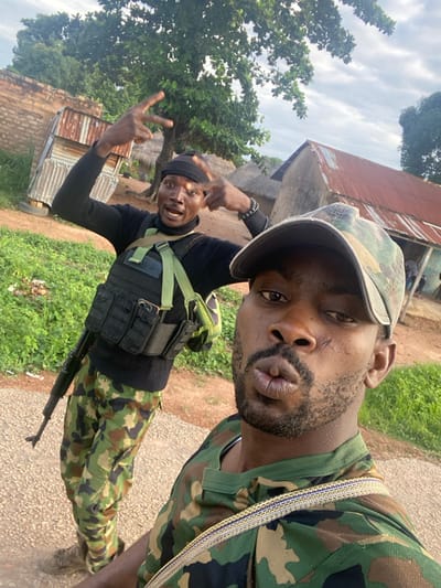 Two adult males are depicted outdoors, in what appears to be a rural or semi-urban setting within Ayati, Nigeria. The image is a selfie taken by the individual in the foreground, who is a dark-skinned male with a beard and a distinctive scar on his left cheek. He wears a camouflage baseball cap and a camouflage short-sleeved shirt, pouting his lips towards the camera.
Behind him, slightly to the left, stands a second dark-skinned male. This individual wears a black beanie, a black long-sleeved turtleneck shirt, camouflage trousers, and a black tactical vest with multiple pouches and light green webbing straps. An assault rifle, appearing to be an AK-pattern variant, is slung across his body. He is posing for the camera with both hands raised, forming "V" (peace) gestures with his fingers, and has his mouth open. A dark beaded bracelet is visible on his left wrist.
The background features simple structures, including one with a rusty red corrugated metal roof and mud-brick walls, and another with silver corrugated metal. Trees with green foliage are visible, along with sparse ground vegetation. The ground consists of bare earth or dirt. The sky is overcast.