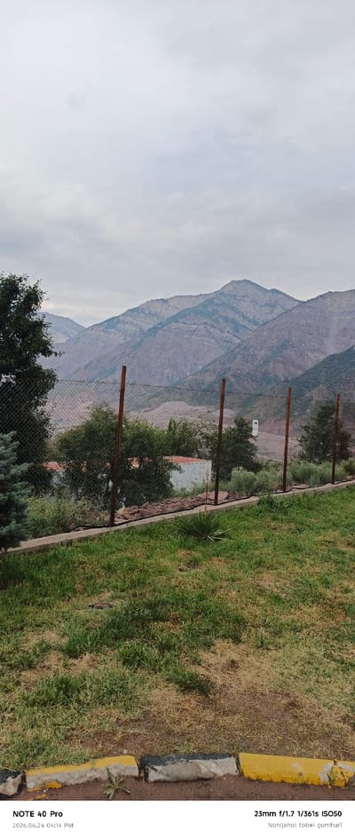 This is a landscape photograph taken in Roghun, Tajikistan. The image captures a view of rugged, mountainous terrain under a cloudy sky. In the foreground, there is a patch of grass with some dried brown areas, bordered by a curb with yellow and black paint. A chain-link fence runs across the middle ground, with wooden posts supporting it. Behind the fence, there are several trees and some small structures with white walls and reddish roofs. The distant mountains display layered rock formations and sparse vegetation. The overall mood is serene, with the overcast sky suggesting a calm, possibly cool, weather condition. The visible text at the bottom indicates the device used (NOTE 40 Pro), the date and time of the photo (2026.06.24 04:14 PM), and camera settings (23mm f/1.7 1/361s ISO50), along with some text in what appears to be Turkish or a similar language. No people are visible in the frame.