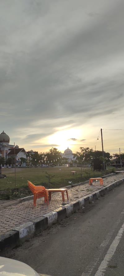 The image depicts an outdoor scene with two prominent domed buildings resembling mosques in the background, one on the left with intricate detailing and another further back on the right. The sky is overcast with clouds, but the setting sun creates a warm glow, indicating it is either sunrise or sunset. The foreground features a paved walkway with a concrete curb separating it from a dirt and asphalt road.
On the walkway, several orange plastic chairs are stacked on top of an orange plastic table. Further down the walkway, another orange plastic table is visible. A few trees are scattered throughout the grassy area between the walkway and the buildings. In the distance, a utility pole with wires is present, and some string lights are hung between trees in front of the buildings. A car is parked on the left side of the grass area.
The scene appears to be a public space, possibly a park or a plaza adjacent to the religious buildings. The time of day is suggested by the low sun angle and the warm light filtering through the clouds. The overall mood is serene, with the empty seating suggesting a quiet moment or the end of a day. No people are visible in the frame. There is no visible text in the image. The location context provided is Meunasahaluedrienlhoksukon Barat, Indonesia, which aligns with the architectural style of the buildings and the tropical vegetation.