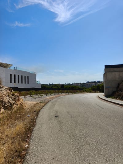 This image depicts an outdoor scene with a curving asphalt road in the foreground. To the left of the road, there is a rocky, dry, and grassy embankment with some scattered litter. Further back on the left, a modern, white building with vertical window slits and solar panels on the roof is visible. In the background, a line of green trees separates the foreground from a residential area with buildings on a hillside, and a construction crane is visible among them. To the right of the road, a concrete structure, possibly part of a building or infrastructure, shows some graffiti. The sky is a clear blue with wispy white clouds, suggesting a sunny day. The overall scene appears to be in a somewhat undeveloped or developing area, likely a suburban or rural hillside. The time of day is likely daytime, given the bright sunlight and clear sky. There are no people visible in the image. The location context provided is Dehesa De Campoamor, Spain.