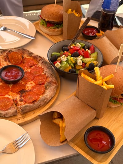 The image shows a table laden with food, suggesting a meal is being served outdoors. The subjects are various food items including a pepperoni pizza, a Greek salad, french fries, and burgers. There are also drinks in bottles and small bowls of ketchup. The setting appears to be a restaurant patio or a backyard, given the casual arrangement of the food and the outdoor lighting.
The activity taking place is eating, or preparing to eat. The food is arranged in a way that suggests a shared meal or a buffet-style service. The lighting indicates it is daytime, possibly late afternoon or early evening, with natural sunlight illuminating the scene. There are no people visible in the image.
Notable details include the rustic presentation of the food, with kraft paper used for the french fries containers and wooden trays for some of the dishes. The pizza has a thick, slightly charred crust, and the Greek salad contains tomatoes, cucumbers, olives, and feta cheese. The french fries are golden brown and appear to be freshly cooked. The ketchup is a vibrant red. There is a bottle of what appears to be a dark beverage, possibly soda or iced tea, with blue and white labeling. The word "SPECIAL" is visible on the bottle label.
The location context is Ptghni, Armenia. While there are no explicit visual cues confirming this location, the overall aesthetic of a relaxed outdoor meal is consistent with a Mediterranean or Eastern European setting. The weather appears to be pleasant, conducive to outdoor dining. There are no strong emotional cues, but the abundance and variety of food suggest a festive or enjoyable occasion.