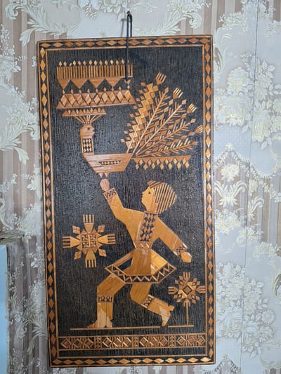 The image shows a straw-crafted wall hanging featuring a stylized human figure carrying a large bird with an elaborate headdress and fanned tail. The figure is depicted in a dynamic pose, seemingly walking or dancing, adorned in clothing with geometric patterns. Two star-like ornaments are positioned on either side of the figure. The artwork is framed with a border of woven straw and mounted against a backdrop of patterned wallpaper. The wall hanging is suspended by a dark cord. The overall scene is an interior setting.