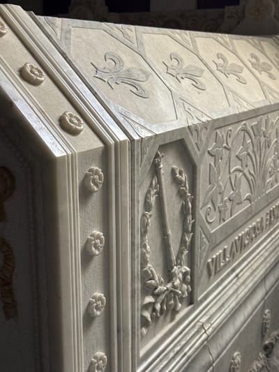 The image shows a close-up, angled view of a marble sarcophagus, likely a tomb, located in the Pantheon of Kings within the Monastery of El Escorial in San Lorenzo de El Escorial, Spain. The sarcophagus features intricate carvings, including floral motifs and what appear to be fleur-de-lis symbols on its lid. The side of the sarcophagus displays a relief carving of a wreath encircling a fasces, a symbol of authority. Partially visible text is etched into the marble on the side. The material is a light-colored marble, and the lighting highlights the detailed stonework. No people are visible in the image.