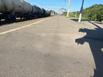 This is a ground-level, wide-angle photograph taken on a sunny day at a train station in Kalynivka, Ukraine. The foreground is dominated by the coarse texture of a paved platform, with a faint yellow line marking its edge. To the left, a long train composed of multiple tank cars is stopped on the tracks. The train cars are light-colored with dark green bands and display various markings and numbers, including "106" and "739 99716". In the mid-ground, the train tracks run parallel to the platform, bordered by sparse green grass. Beyond the tracks, a row of utility poles with overhead electrical wires for the trains is visible, leading into a distant treeline of lush green trees. A small, open-air station shelter with a curved roof stands further down the platform, where a single person is seated. The sky is a clear, bright blue. The right side of the image features the shadow of a person, possibly the photographer, wearing a hat, cast across the platform.
