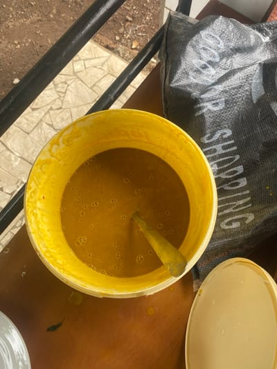 The image shows a yellow bucket filled with a thick, yellow liquid, possibly a sauce or soup, with a metal spoon inside. The bucket is sitting on a wooden table, and part of a black plastic bag with white lettering is visible in the upper right corner of the image. The table is outside, as can be seen by the ground and a railing in the background. The coordinates provided correspond to a location in Limbe, Cameroon, but the image does not provide any contextual information about the location. There are no people in the image.