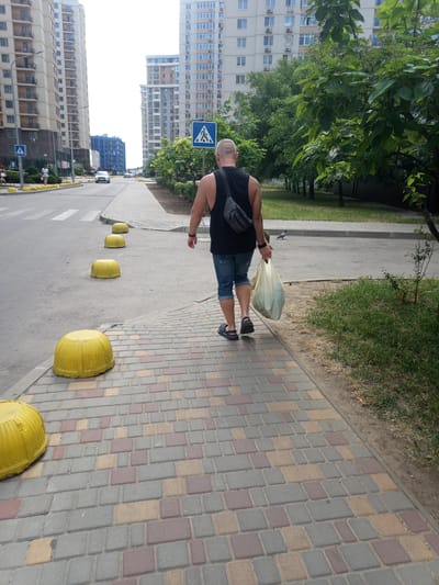 A man with a shaved head, wearing a black tank top, denim shorts, and a dark crossbody bag, is walking away from the camera on a paved sidewalk. He is carrying a translucent plastic bag filled with items in his right hand. The sidewalk is made of multi-colored rectangular pavers. Yellow, dome-shaped bollards are spaced along the edge of the sidewalk, separating it from a street. The street is asphalt and has white pedestrian crossing markings. On the left side of the street, tall residential buildings with numerous windows are visible. On the right side of the street, beyond a grassy area, are more trees and buildings. A pedestrian crossing sign is visible on a pole to the left of the man. The sky is overcast. The scene appears to be in a residential area of the city of Lymanka, Ukraine.